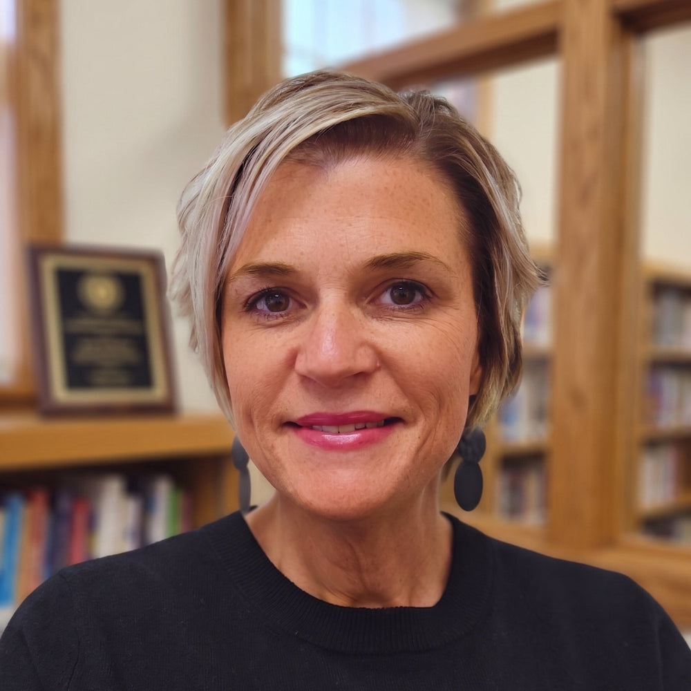 A woman with short blonde hair wearing a black crewneck sweater and dark earrings, standing in front of a wooden bookshelf.